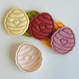 Pu&ograve; includere: Quattro stampini per biscotti colorati a forma di favo con un cuore al centro. Gli stampini per biscotti sono disposti in cerchio con uno stampino per biscotti bianco al centro.
