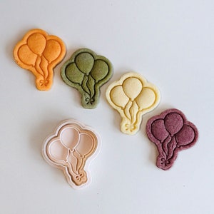 May include: Five cookie cutters in the shape of a bunch of balloons. The cutters are in various colors, including orange, green, yellow, cream, and purple.