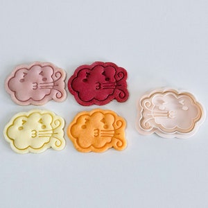 May include: Five cookie cutters in the shape of a cloud with a face blowing air. The cutters are in various colors: pink, red, yellow, orange, and white.