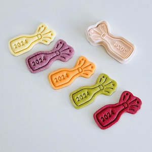 May include: Five colourful cookie cutters shaped like champagne bottles with the year 2024 embossed on them. The cookie cutters are arranged in a row on a light grey surface.