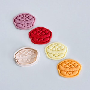 May include: Five colourful cookie cutters shaped like pies with smiling faces. The cutters are pink, red, beige, yellow, and orange.