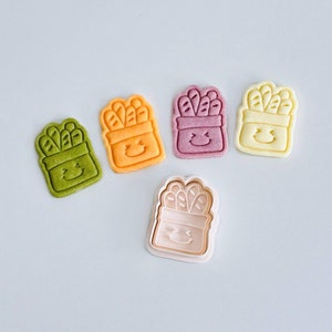 Op de afbeelding: Vijf koekjesvormen in verschillende kleuren, gevormd als een container met een glimlachend gezicht en een bosje broodstokjes erin. De koekjesvormen staan op een lichtblauwe achtergrond.