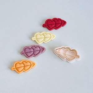 May include: An assortment of heart-shaped cookie cutters in various colors, including red, yellow, purple, orange, and pink. Each cutter features two connected hearts. The cutters are arranged on a light background.
