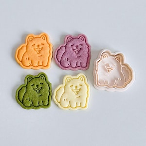 Puede incluir: Un surtido de galletas con forma de perro en varios colores: naranja, morado, verde y amarillo. También hay un cortador de galletas rosa. Las galletas tienen una cara sonriente y un aspecto esponjoso, perfectas para un dulce temático.