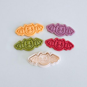 Puede incluir: Cinco cortadores de galletas con forma de murciélago, cuatro son de masa coloreada, uno es de plástico blanco. Los cortadores están dispuestos en círculo con el cortador de plástico en el centro.