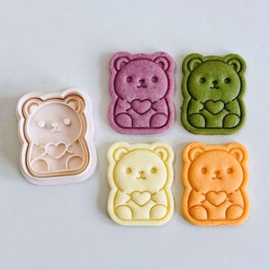 Può includere: Una collezione di formine per biscotti e biscotti a forma di orsetto. I biscotti sono nelle tonalità del viola, verde, giallo e arancione, ognuno con un dettaglio a cuore. La formina è rosa chiaro.