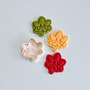 May include: Four cookie cutters in the shape of a maple leaf with a cute face. The cutters are green, orange, white, and red.