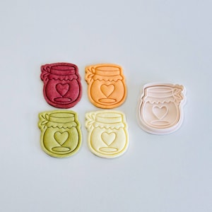 May include: Five cookie cutters in the shape of a jar with a heart on the front. The cutters are in various colors: red, orange, green, yellow, and white.