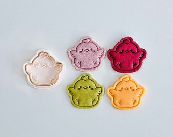 May include: A collection of six chick-shaped items. One is a light pink cookie cutter, and the others are cookies in shades of pink, red, green, and orange. Each chick has a heart on its head and small wings.