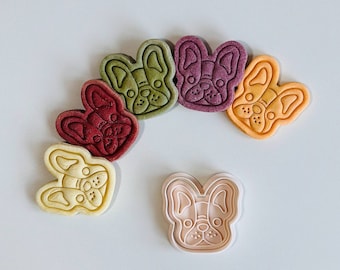May include: A set of six dog-shaped cookie cutters in various colours, including green, purple, orange, red, yellow, and pink. The cutters are arranged in a semi-circle with the pink cutter in the centre.