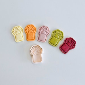 Könnte beinhalten: Sechs Ausstecher in Form von Pfotenabdrücken in verschiedenen Farben, darunter Gelb, Orange, Rosa, Grün und Rot. Die Ausstecher sind in einem Halbkreis angeordnet, mit einem weißen Pfotenabdruck-Ausstecher in der Mitte.