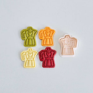 Può includere: Cinque stampini per biscotti a forma di caffettiera con un cuore sul lato. Gli stampini sono di vari colori: verde, arancione, beige, rosso e bianco.