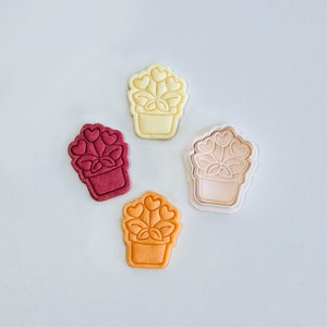 Puede incluir: Cuatro cortadores de galletas con forma de planta en maceta con flores en forma de corazón. Los cortadores son de diferentes colores: blanco, rojo, naranja y rosa.