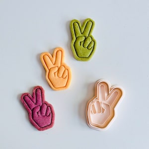 Può includere: Quattro stampini per biscotti a forma di segno della pace, uno è di plastica bianca, gli altri sono verde, giallo e viola.