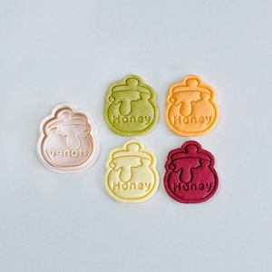 Może przedstawiać: Pięć foremek do ciasteczek w kształcie słoika miodu, jedna zielona, jedna pomarańczowa, jedna żółta, jedna czerwona i jedna biała. Formy znajdują się na białym tle. Na każdej formie widnieje napis "Honey".