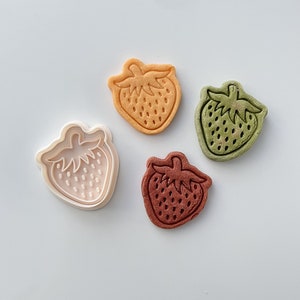 May include: Three strawberry-shaped cookie cutters, one white, one orange, and one green, with a brown strawberry-shaped cookie on a white background.