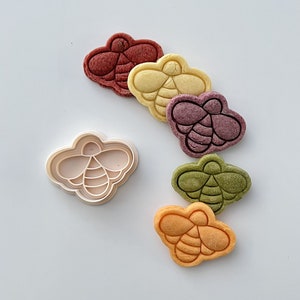 May include: A white plastic bee-shaped cookie cutter with five colorful bee-shaped cookies. The cookies are red, yellow, purple, green, and orange.
