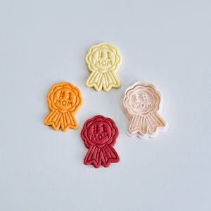May include: Four cookie cutters in the shape of a ribbon with the text "#1 MOM" on them. The cutters are in different colours: yellow, orange, red, and beige.