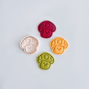 May include: Four cookie cutters in the shape of mushrooms with smiling faces. The cutters are red, orange, green, and white.