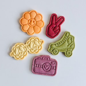 Może przedstawiać: Pięć kolorowych ciasteczek w kształcie kwiatu, znaku pokoju, aparatu fotograficznego, rolek i gestu dłoni z symbolem pokoju.