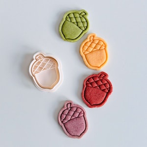 May include: An assortment of acorn-shaped cookies in various colors, including green, orange, red, and purple. One cookie cutter is also visible. The cookies have a detailed design on the top half, and are arranged on a white surface.