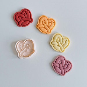 Puede incluir: Cinco cortadores de galletas con forma de pretzel, con una cara sonriente. Los cortadores son de varios colores, incluyendo rojo, naranja, amarillo y rosa.