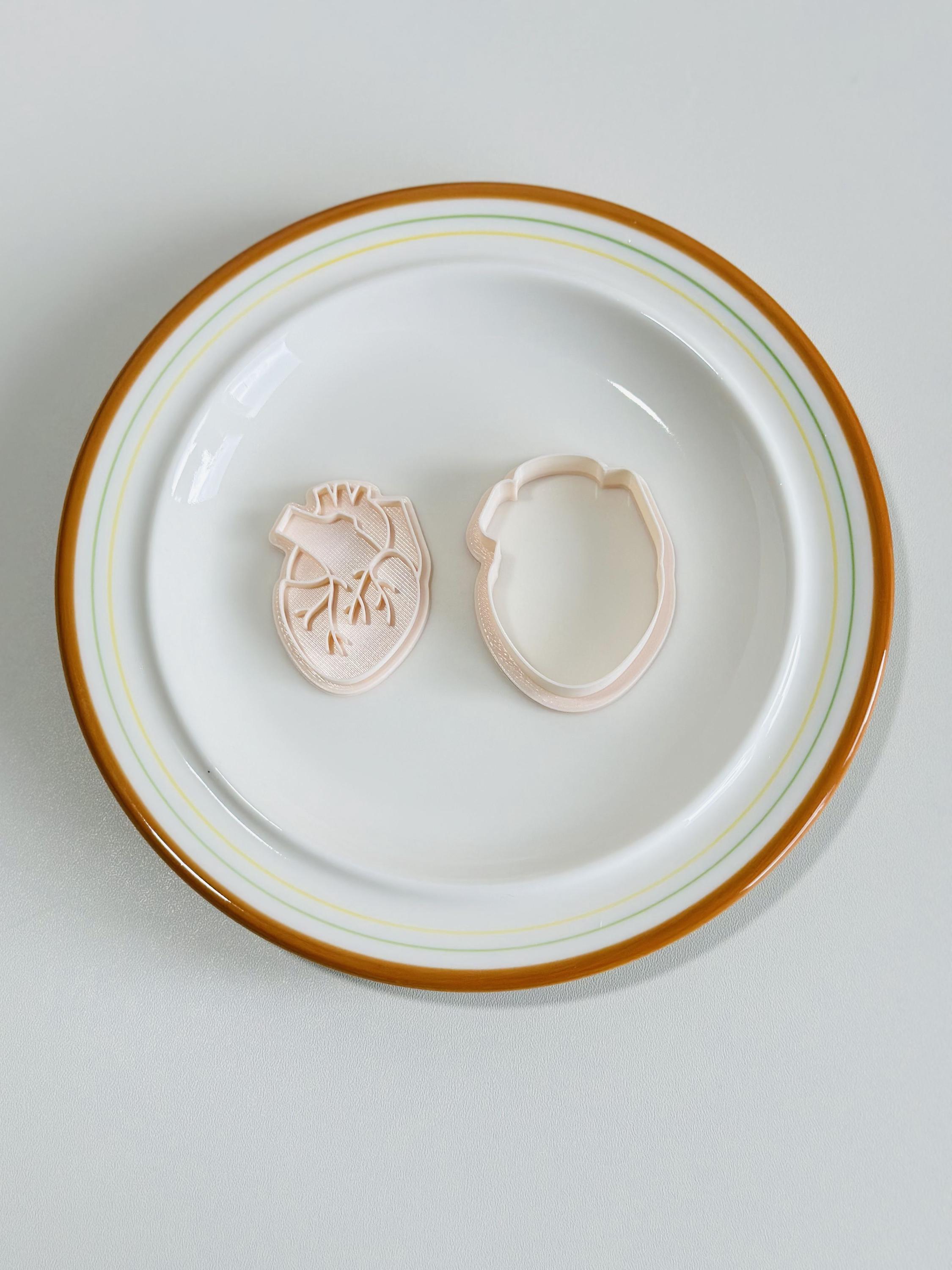 May include: Two plastic cookie cutters in the shape of a human heart, one with the internal anatomy of the heart visible, and the other a solid outline. The cutters are on a white plate with a brown rim.