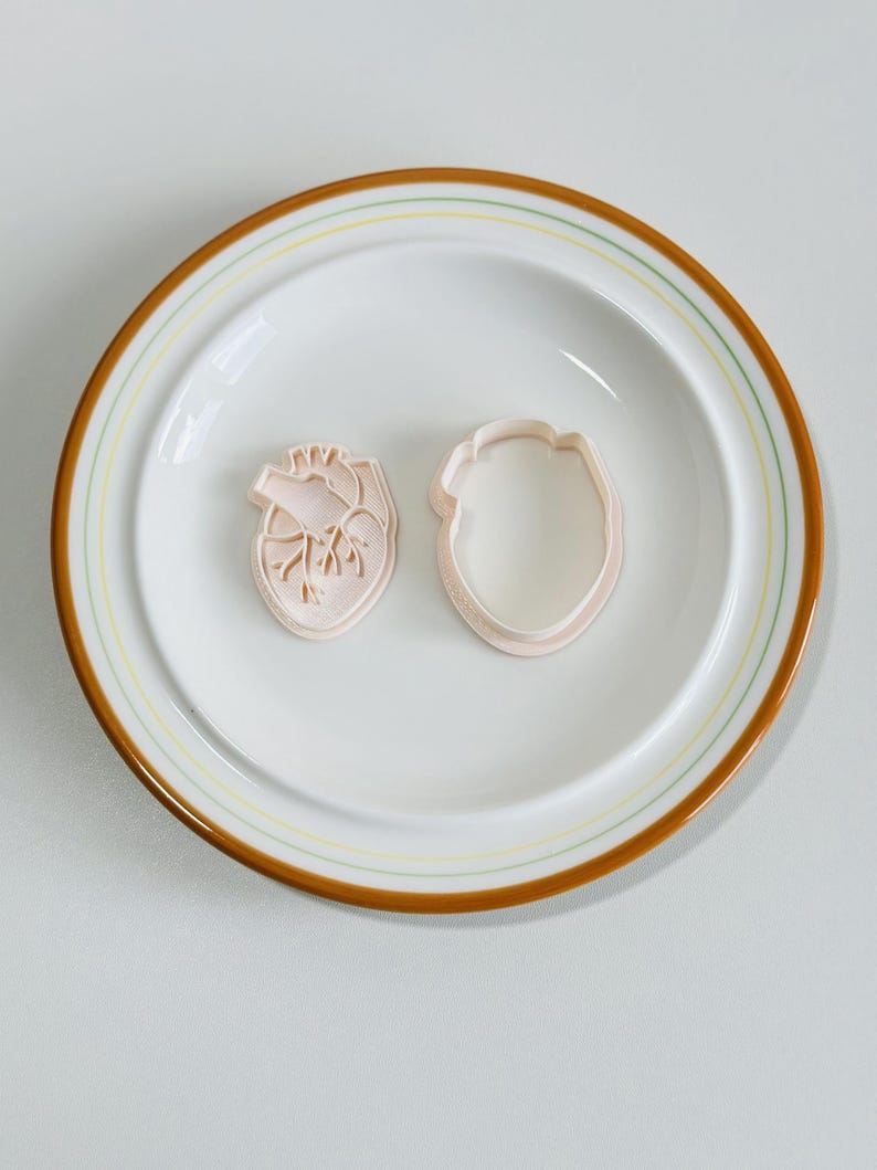 May include: Two plastic cookie cutters in the shape of a human heart, one with the internal anatomy of the heart visible, and the other a solid outline. The cutters are on a white plate with a brown rim.