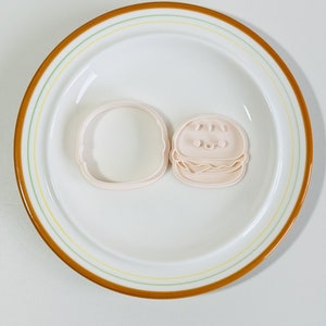 Puede incluir: Dos cortadores de galletas de pl&aacute;stico rosa con forma de hamburguesa. El cortador superior tiene un pan de s&eacute;samo, dos ojos y una sonrisa. El cortador inferior es un pan simple.