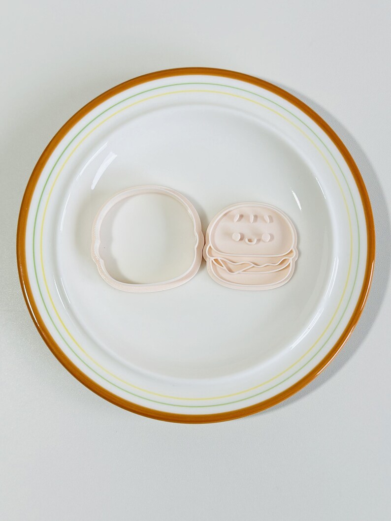 Puede incluir: Dos cortadores de galletas de pl&aacute;stico rosa con forma de hamburguesa. El cortador superior tiene un pan de s&eacute;samo, dos ojos y una sonrisa. El cortador inferior es un pan simple.