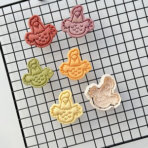 May include: Six colorful cookie cutters shaped like chickens on a black wire cooling rack. The cutters are made of plastic and have a detailed design.
