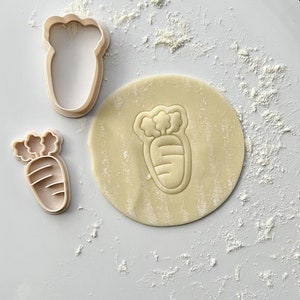 May include: Two plastic cookie cutters in the shape of a carrot. The larger cutter is on a piece of dough, and the smaller cutter is next to it. The dough is covered in white flour.