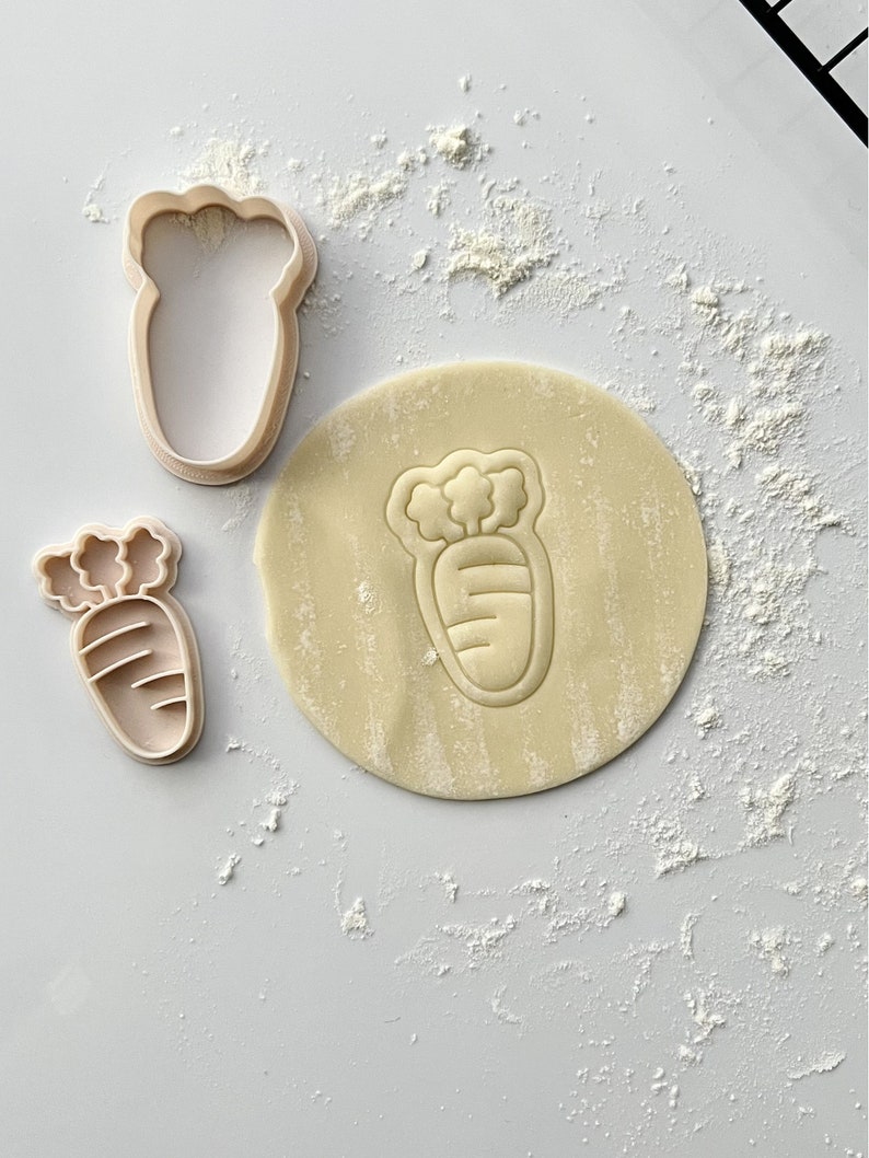 May include: Two plastic cookie cutters in the shape of a carrot. The larger cutter is on a piece of dough, and the smaller cutter is next to it. The dough is covered in white flour.