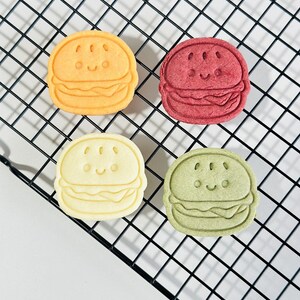 Puede incluir: Cuatro galletas con forma de hamburguesa con caras de dibujos animados, de colores amarillo, rojo, blanco y verde, sobre una rejilla de alambre negra.