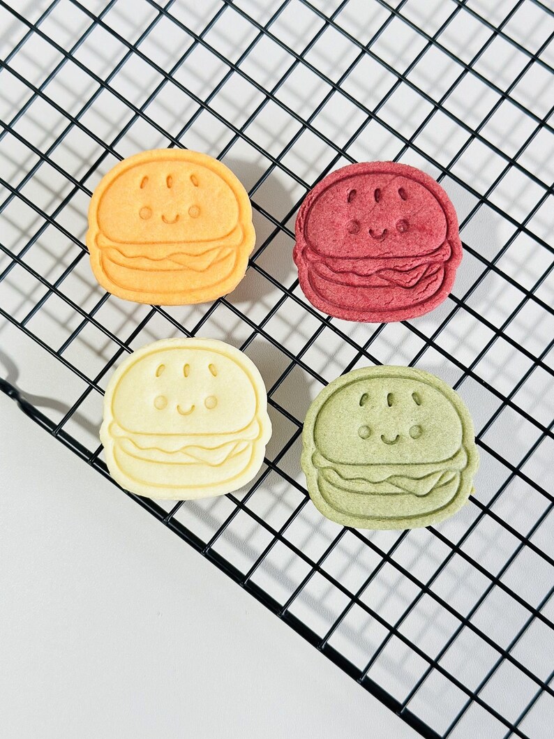 Puede incluir: Cuatro galletas con forma de hamburguesa con caras de dibujos animados, de colores amarillo, rojo, blanco y verde, sobre una rejilla de alambre negra.