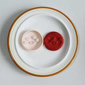 May include: A white plate with a brown and yellow rim holds a light pink cookie cutter and a red cookie. The cookie cutter has a smiley face and heart shapes. The red cookie has a smiley face.