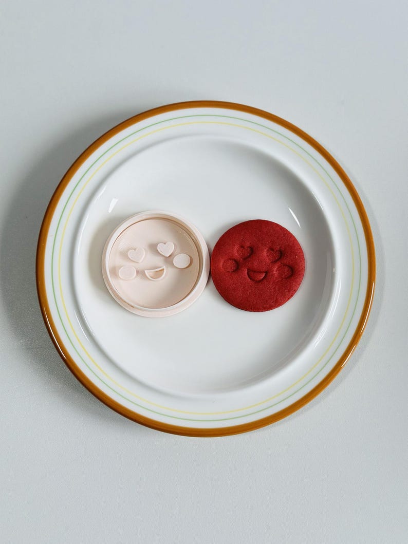 May include: A white plate with a brown and yellow rim holds a light pink cookie cutter and a red cookie. The cookie cutter has a smiley face and heart shapes. The red cookie has a smiley face.