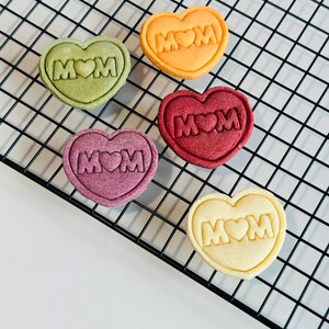 May include: Five heart-shaped cookies on a black wire rack. The cookies are decorated with the word "MOM" and a heart. The cookies are in different colors: green, orange, red, purple, and white.