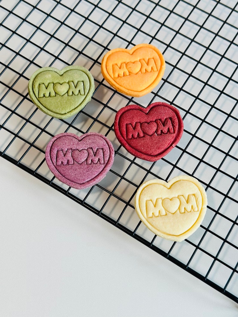 May include: Five heart-shaped cookies on a black wire rack. The cookies are decorated with the word "MOM" and a heart. The cookies are in different colors: green, orange, red, purple, and white.