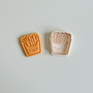 May include: A white plastic cookie cutter shaped like a cartoon french fry container with a smiling face. Next to it is a brown cookie cut out in the same shape.