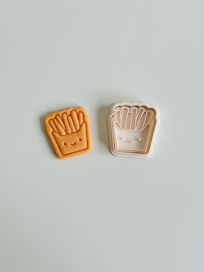 May include: A white plastic cookie cutter shaped like a cartoon french fry container with a smiling face. Next to it is a brown cookie cut out in the same shape.
