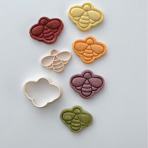 May include: A set of six bee-shaped cookie cutters, including one cloud-shaped cutter. The cutters are made of white plastic and are arranged in a row on a white background. The bee cutters are in various colors, including yellow, orange, red, purple, and green.