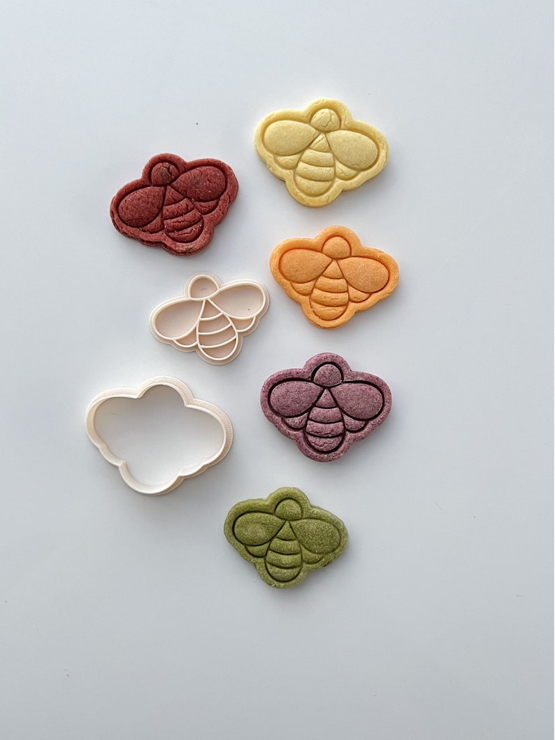 May include: A set of six bee-shaped cookie cutters, including one cloud-shaped cutter. The cutters are made of white plastic and are arranged in a row on a white background. The bee cutters are in various colors, including yellow, orange, red, purple, and green.