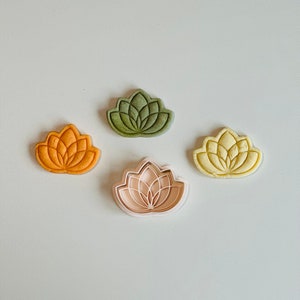 May include: Four cookie cutters in the shape of a lotus flower. One cutter is green, one is yellow, one is orange, and one is pink.