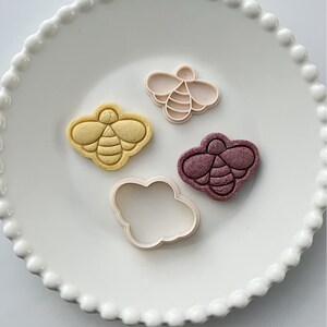 May include: Four cookie cutters in the shape of a bee. Two are white, one is yellow, and one is purple. The cutters are on a white plate with a beaded edge.