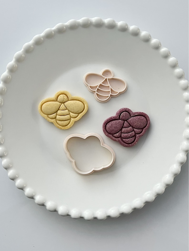May include: Four cookie cutters in the shape of a bee. Two are white, one is yellow, and one is purple. The cutters are on a white plate with a beaded edge.