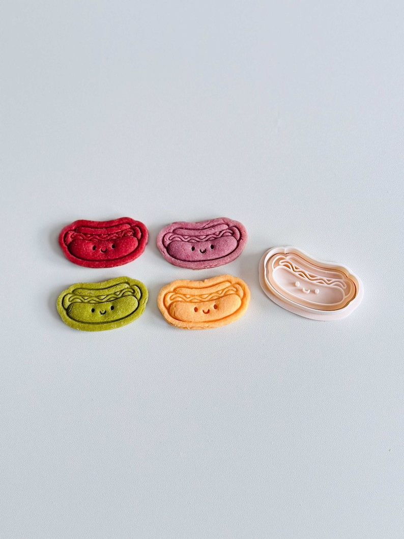 May include: Four colorful cookie cutters shaped like hot dogs with smiling faces. The cutters are arranged in a row, with a fifth cutter in the center, which is a larger version of the hot dog cutter.