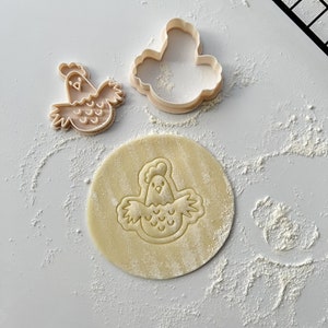 May include: Two plastic cookie cutters in the shape of a chicken, one with a solid outline and the other with a detailed design. A round piece of dough is imprinted with the detailed chicken design.