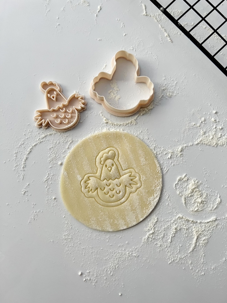 May include: Two plastic cookie cutters in the shape of a chicken, one with a solid outline and the other with a detailed design. A round piece of dough is imprinted with the detailed chicken design.