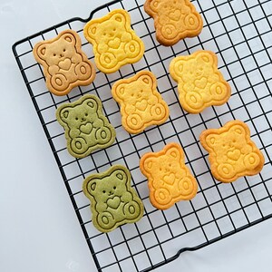 May include: Nine bear-shaped cookies on a black wire cooling rack. The cookies are in three colors: yellow, green, and a light brown. Each cookie has a heart design in the center.